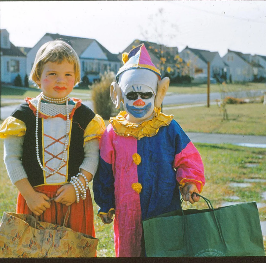 Two young children in Halloween costumes stand outside on a suburban street. One is dressed as a princess with a beaded necklace, and the other as a colorful clown with a painted face and pointy hat. Both hold bags for trick-or-treating.