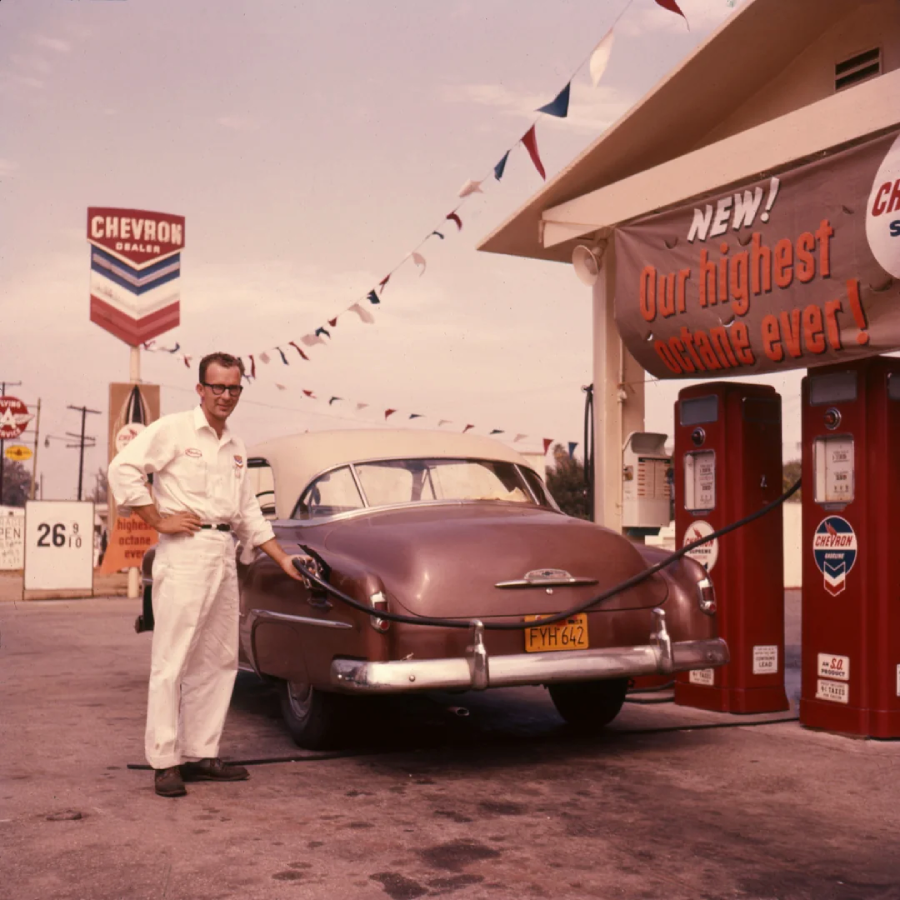 A man in a white uniform pumps gas into a vintage car at a Chevron station. Colorful pennant flags hang above, and a sign advertises high-octane fuel. A gas price sign in the background reads 26.9 cents per gallon.
