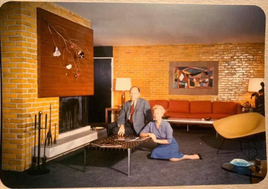 A man in a suit and a woman in a blue dress sit beside a coffee table in a mid-century modern living room with brick walls, a fireplace, a red sofa, abstract art, and warm lighting.