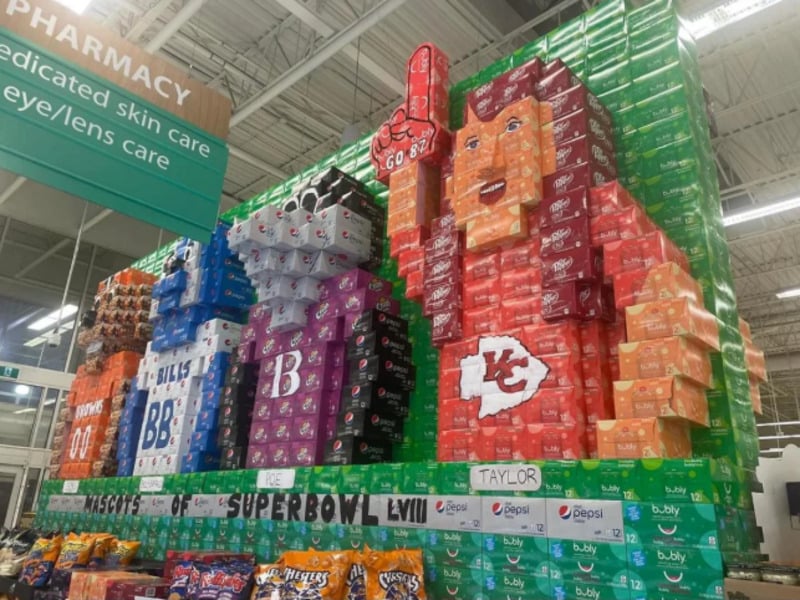 A store display made of soda boxes depicts NFL team jerseys, a foam finger, and a Kansas City Chiefs logo, promoting the Super Bowl. A sign for the pharmacy hangs above the colorful soda box arrangement.