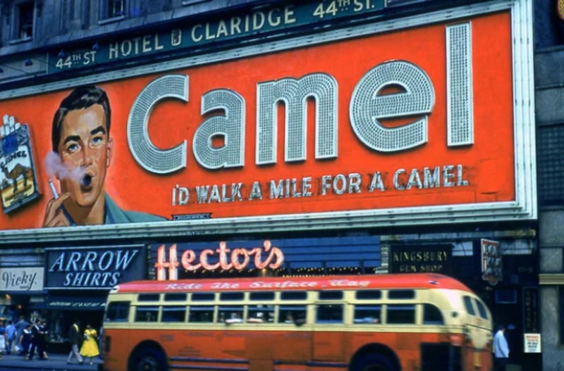 A large vintage Camel cigarette billboard featuring a man smoking and the slogan "I'd walk a mile for a Camel" is displayed above store signs and a red and yellow bus on a busy city street.