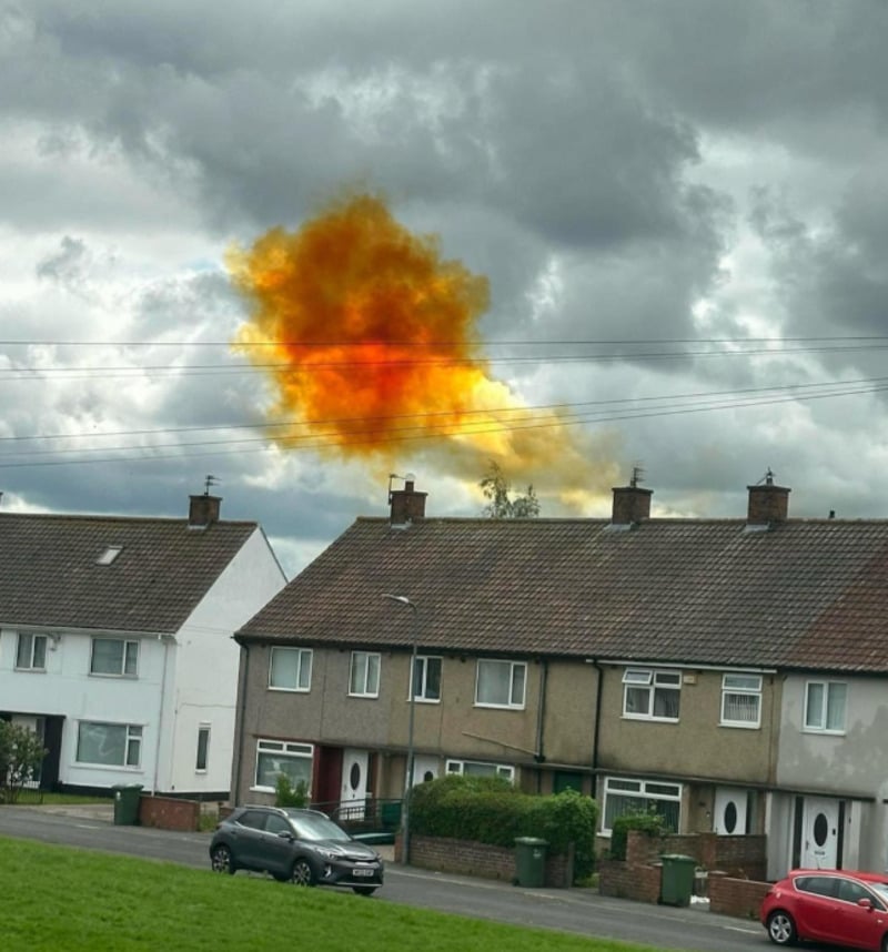 A vivid orange and yellow cloud of smoke or gas hovers above the rooftops of a row of suburban houses on a cloudy day, with several cars parked along the street.