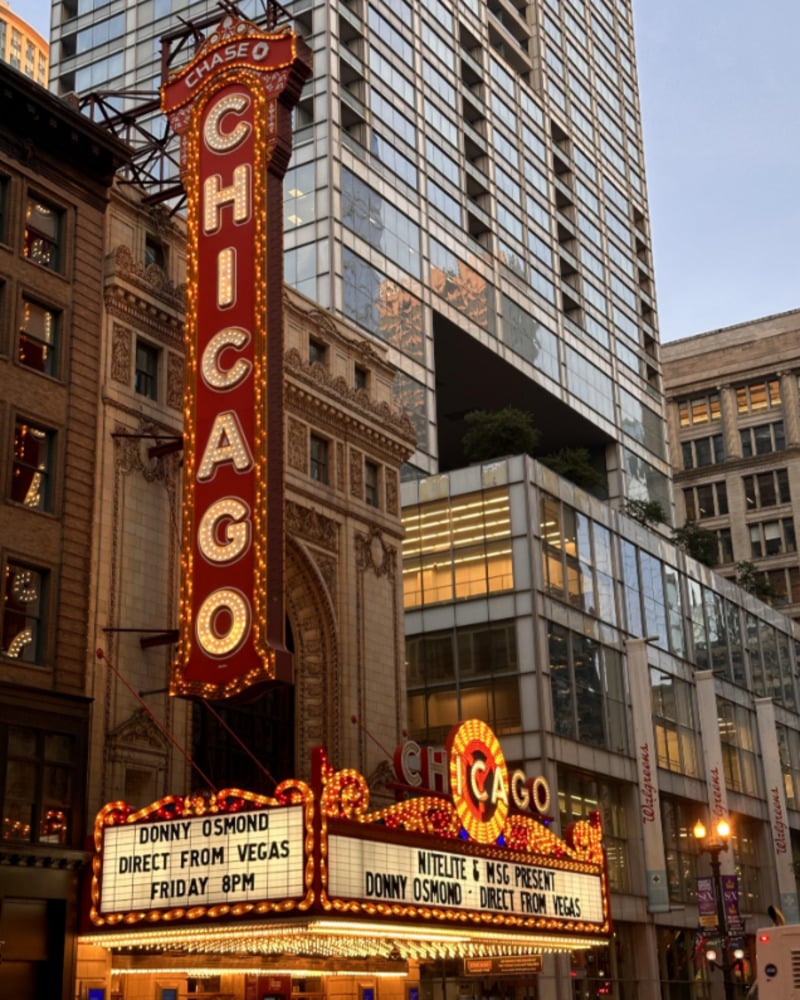 The brightly lit marquee of the Chicago Theatre displays "Donny Osmond Direct from Vegas Friday 8PM" against a backdrop of historic and modern buildings in downtown Chicago.