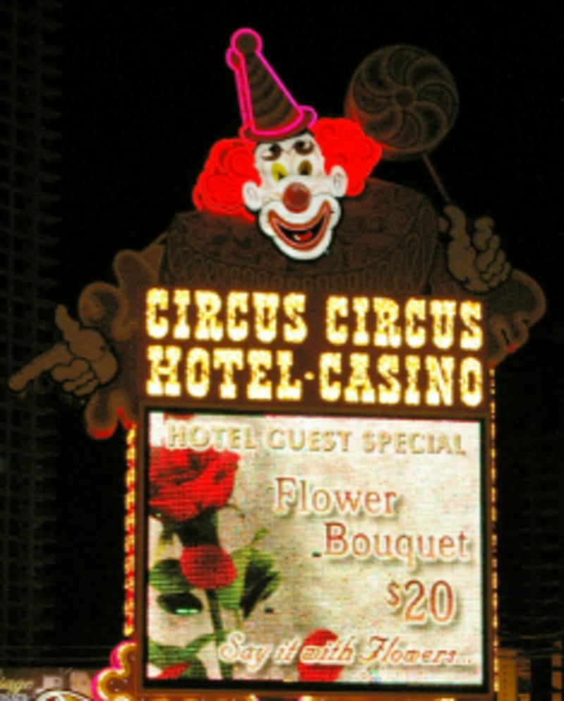 A brightly lit Circus Circus Hotel-Casino sign at night features a large clown holding a lollipop. Below, a digital screen advertises a hotel guest special for a flower bouquet for $20.