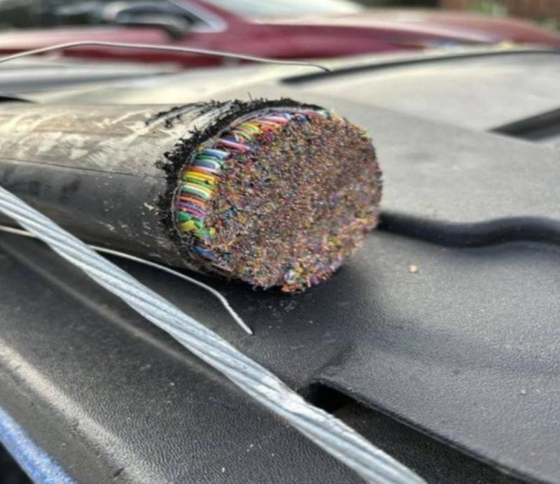 A close-up of a thick black cable cleanly cut open, revealing hundreds of colorful smaller wires tightly packed inside. The cable rests on a dark surface, with a red car blurred in the background.