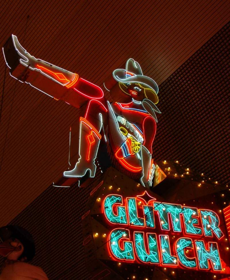A colorful neon sign of a cowgirl in a hat and boots sitting with one leg raised above the words "Glitter Gulch" in bright, illuminated letters. The background is dark, emphasizing the vibrant lights.