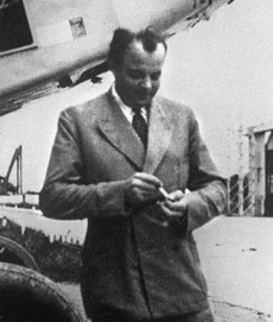 Black and white photo of a man in a suit and tie standing outdoors, smiling and holding a small object in his hands, with a large vehicle or aircraft structure in the background.