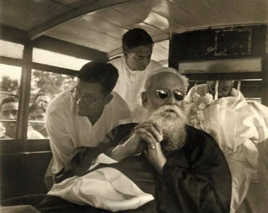 An elderly man with a long white beard and dark sunglasses sits in a vehicle, hands clasped, while two men lean toward him. Several people are visible outside the vehicle in the background.