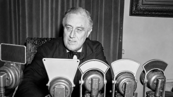 A man in a suit sits at a desk behind several vintage microphones, appearing to speak or prepare for a broadcast. Velvet curtains and a framed picture are visible in the background.