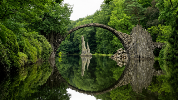 A stone arch bridge spans a calm river, surrounded by lush green trees. The bridge and stone columns reflect perfectly in the water, forming a near-complete circle.