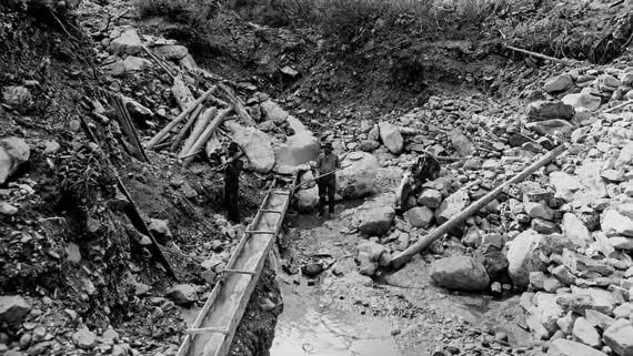 Three people stand in a rocky, muddy trench, working with wooden beams and a makeshift water channel. The area is surrounded by stones, dirt, and sparse vegetation.
