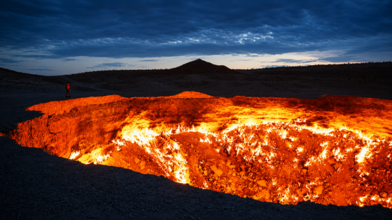 A large fiery crater glows bright orange under a dark, cloudy sky at dusk, with a lone person standing at the edge and a distant hill in the background. earth