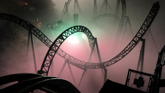 A twisting steel roller coaster illuminated by eerie green and pink lights, with dense fog surrounding the tracks and obscuring the background.