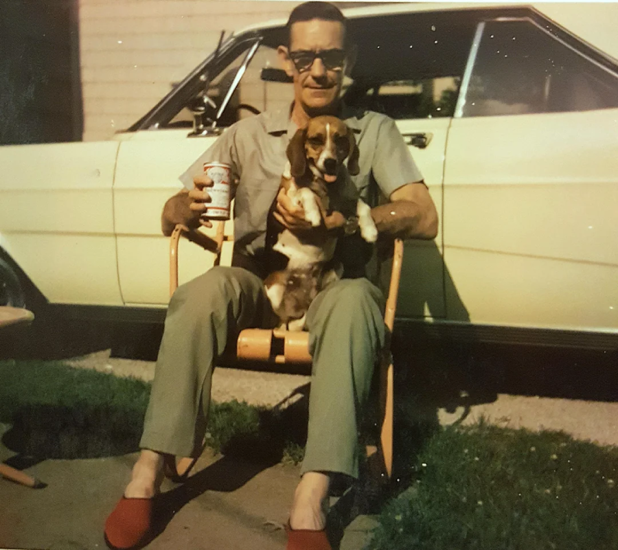 A man wearing sunglasses and red slippers sits in a wooden rocking chair, holding a beagle dog on his lap. He holds a can of beer. A vintage white car is parked behind him. The scene is outdoors.