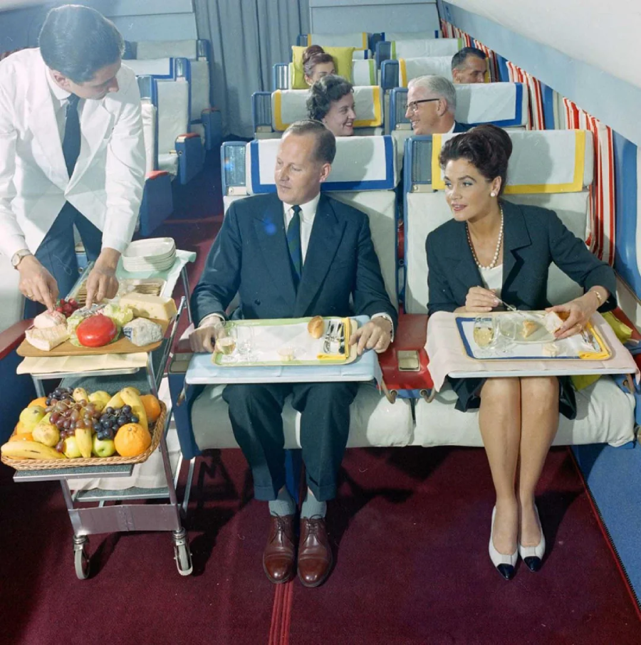 A flight attendant serves a well-dressed man and woman gourmet food from a cart in a vintage airplane cabin with colorful striped seats; other passengers are seated behind them.