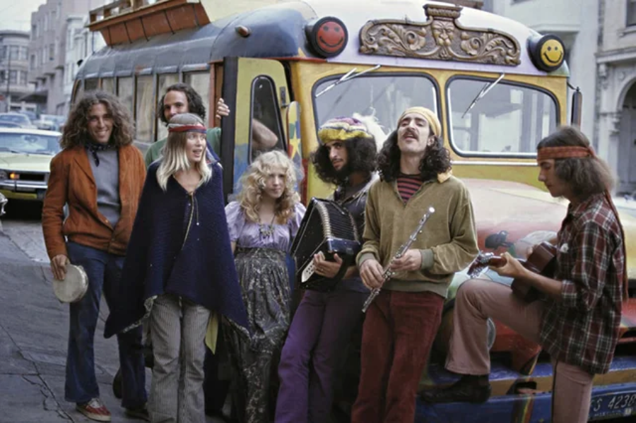 A group of people dressed in colorful, hippie-style clothing stand and sit in front of a brightly painted bus. Some play musical instruments, and everyone appears relaxed and cheerful.