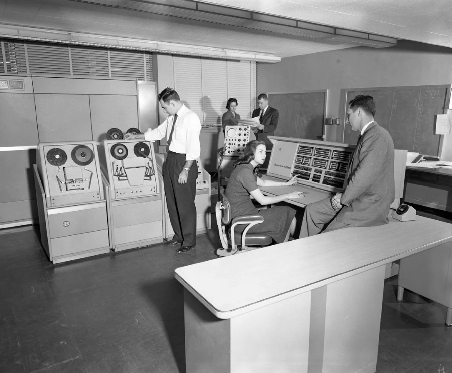 A black-and-white photo of a 1950s computer lab shows five people working with large reel-to-reel computers and control panels. One woman sits at a console while others stand by equipment or talk.