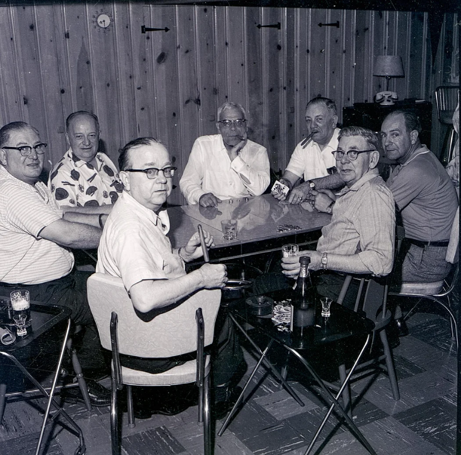 Seven older men sit around a table in a wood-paneled room, holding drinks and playing cards. Some look toward the camera, and drinks and bottles are on the table. The atmosphere appears relaxed and social.