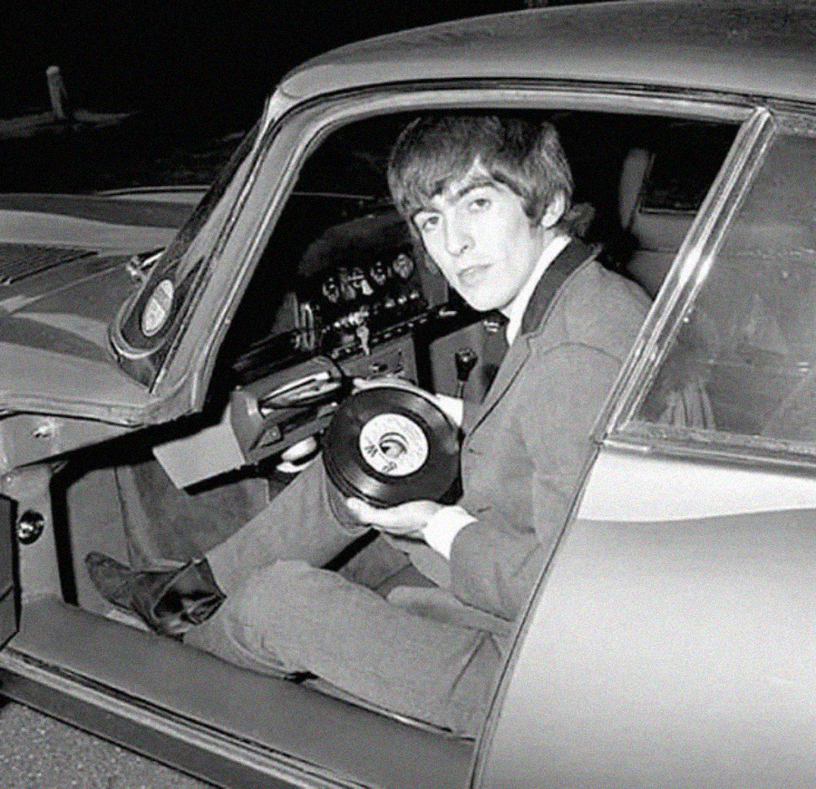 A young man in a suit sits in the driver’s seat of a classic car with the door open, holding vinyl records and looking at the camera. The interior dashboard and steering wheel are visible.