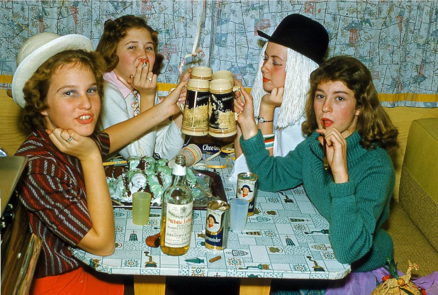 Four teen girls in 1950s-style outfits sit around a table with drinks, snacks, and a bottle of Seagram’s whiskey, playfully posing and pretending to smoke. The setting appears cozy, with vintage decor and patterned curtains.