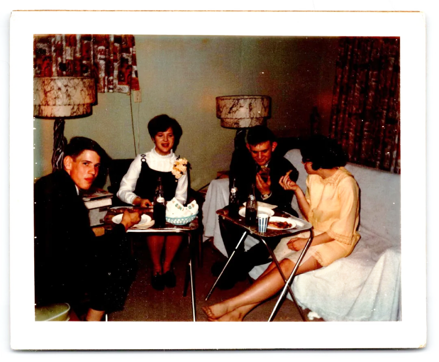 Four adults sit in a cozy living room, eating and drinking around small tray tables. Two women, one in white and one in yellow, and two men in dark suits, chat and share a meal. Lamps and curtains decorate the background.