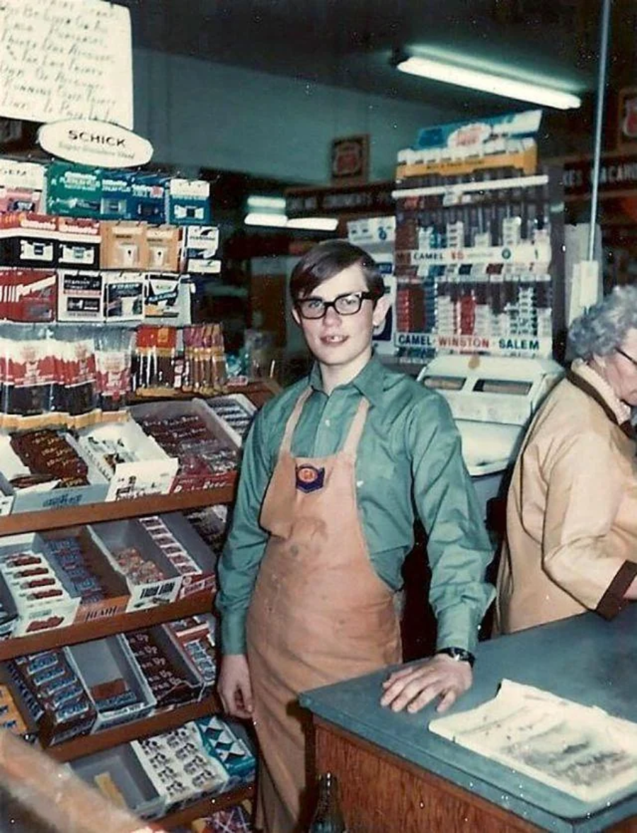 A young man in glasses and a tan apron stands behind the counter of a vintage convenience store, surrounded by shelves stocked with cigarettes, candy, and other goods. An older woman is at the cash register nearby.