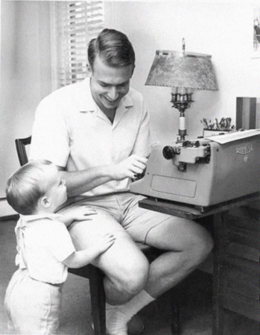 A man in a short-sleeved shirt and shorts types on a vintage typewriter while a small child, standing nearby, looks up at him. They are indoors, near a desk with a lamp and window blinds in the background.