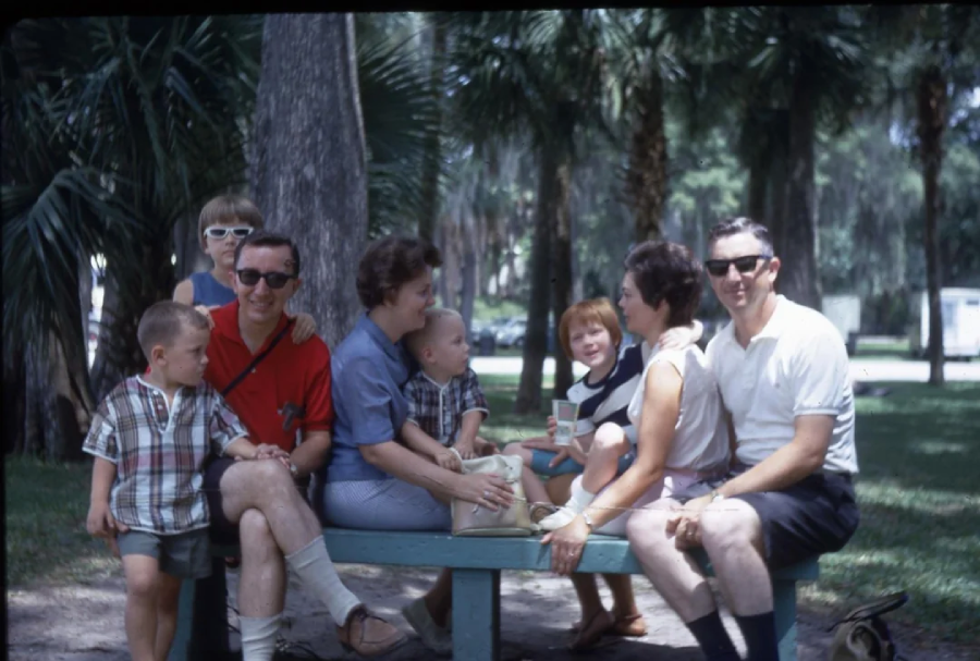 A group of adults and children, casually dressed in summer clothes, sit and chat on a bench in a park surrounded by palm trees. The atmosphere appears relaxed and friendly.