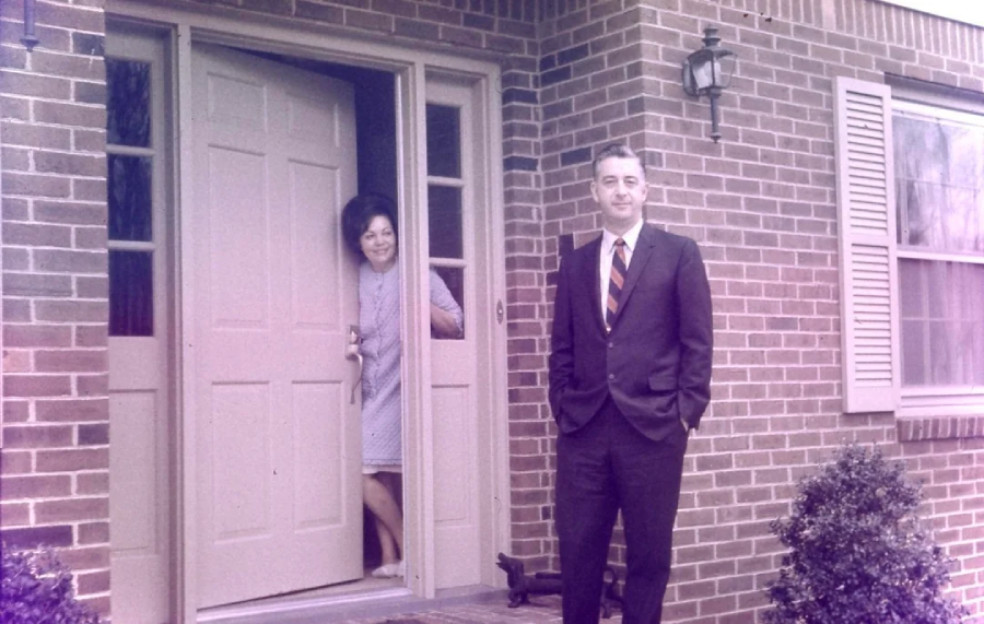 A woman in a light dress stands smiling in the doorway of a brick house, while a man in a suit and striped tie stands outside on the porch, hands in his pockets.