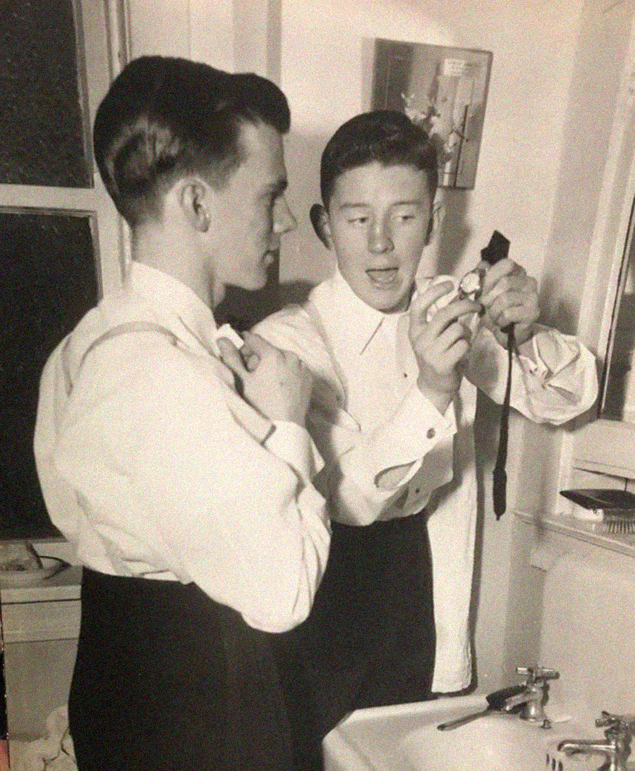 Two young men in dress shirts stand by a bathroom sink; one is holding a bow tie and appears to be demonstrating how to tie it, while the other watches and listens intently. Early-to-mid 20th century setting.