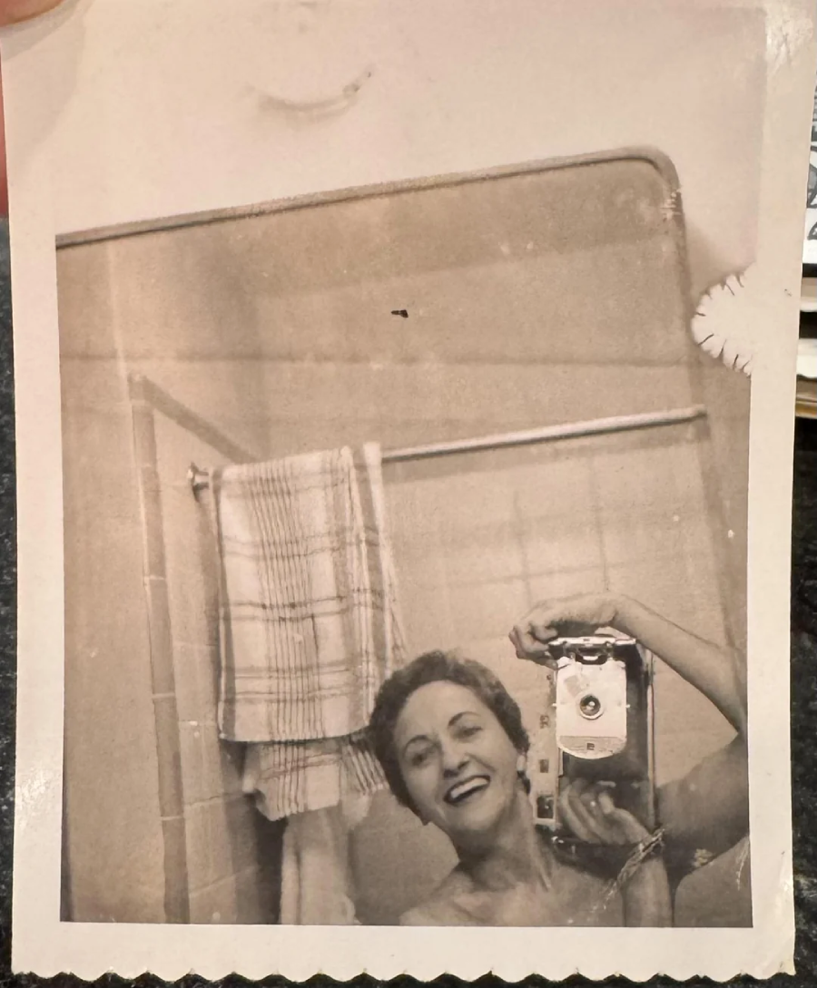 A black-and-white vintage photo of a smiling person taking a mirror selfie in a bathroom, holding a box camera. A towel hangs on a rack behind them. The photo has scalloped edges and signs of wear.