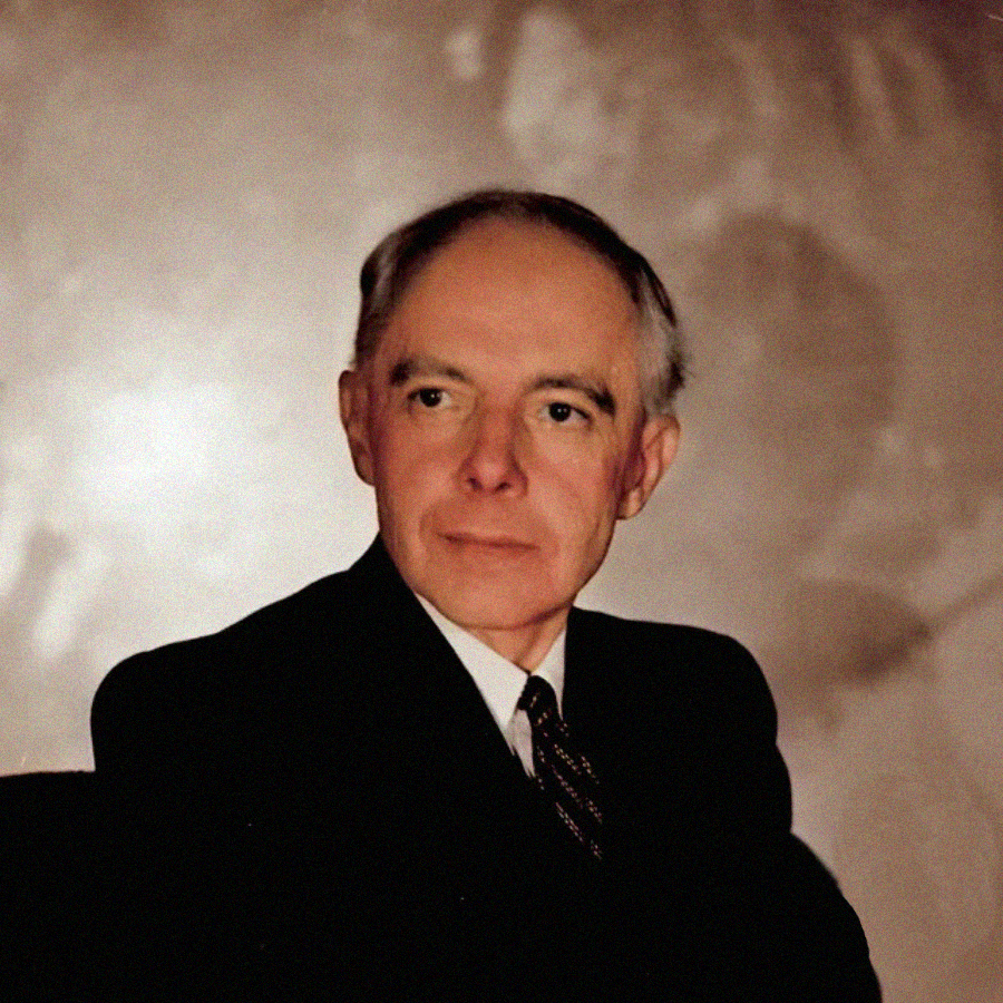 An elderly man wearing a dark suit and tie sits in front of a neutral, softly lit background, looking slightly to his right with a calm expression.