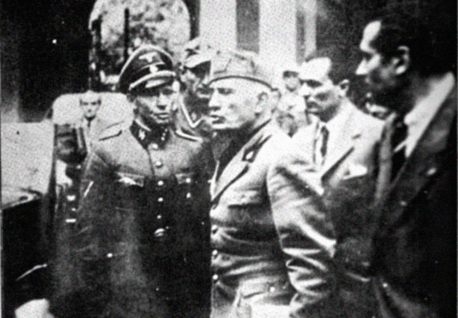 Black-and-white photo of men in military uniforms and suits standing together, having a serious conversation; the scene appears historical, possibly from the World War II era.