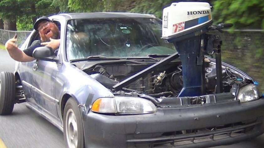 A man leans out of the driver’s window of a car missing its hood, with an outboard boat motor installed in the engine bay. He gestures enthusiastically while driving on a road with trees in the background.