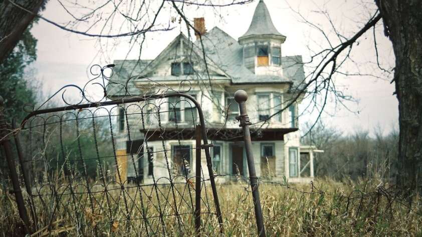 A weathered iron gate stands in tall grass before an old, abandoned Victorian-style house with boarded windows, peeling paint, and a turret, surrounded by bare trees under a cloudy sky.