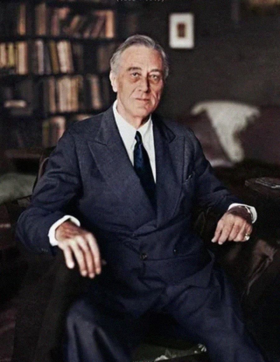 A man in a dark suit sits confidently in an armchair, with a bookshelf and a patterned blanket visible in the background.