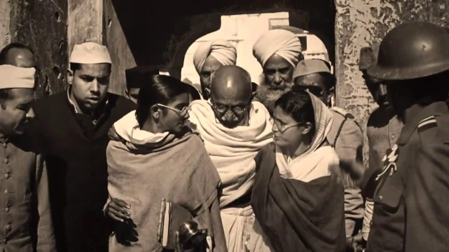 A sepia-toned historical scene shows Mahatma Gandhi surrounded by followers and supporters, some in traditional Indian attire and others in uniforms, as they walk together, possibly during the Indian independence movement.