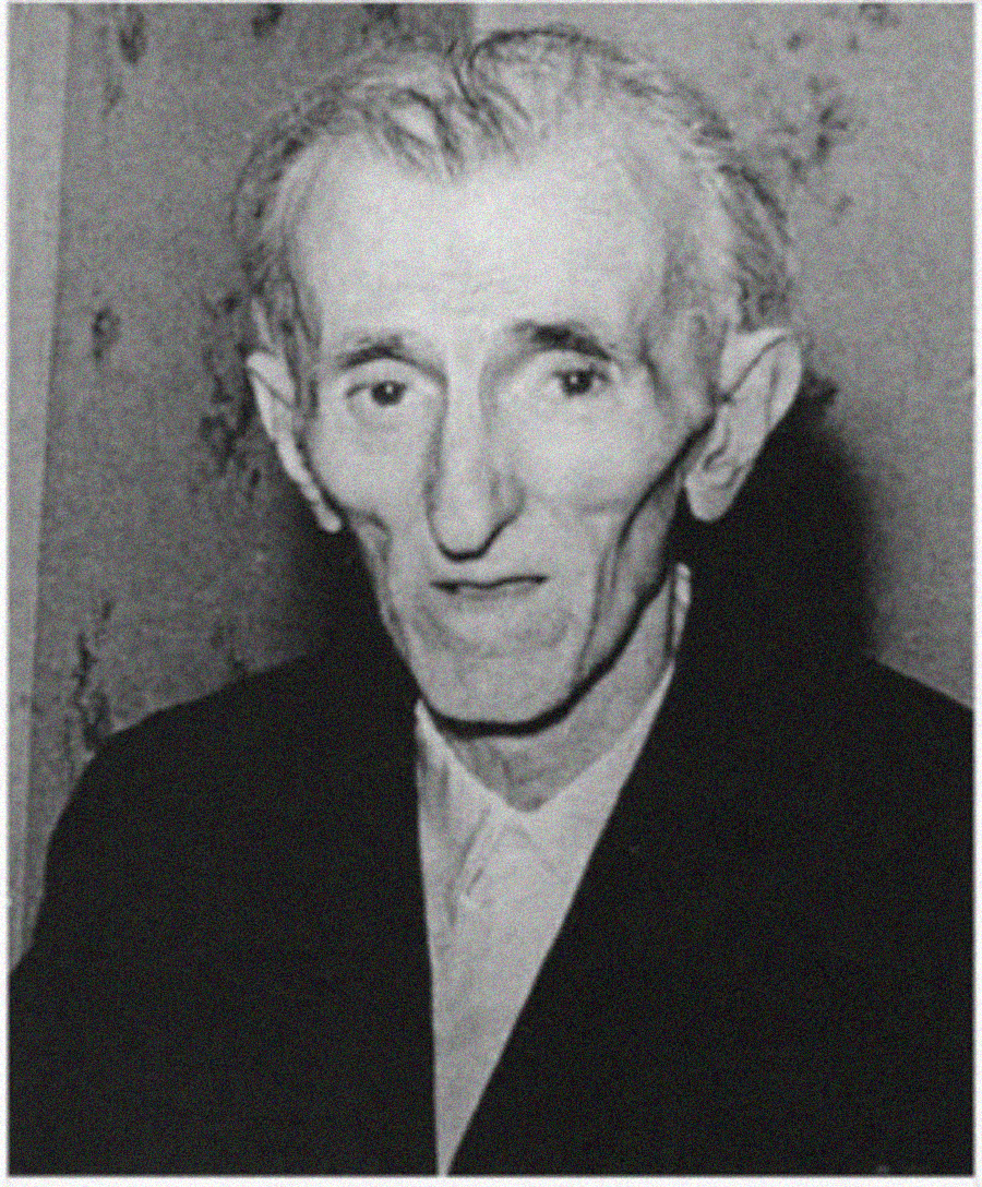 An elderly man with thin face, prominent cheekbones, and swept-back hair, wearing a dark jacket and light shirt, looks toward the camera in a black and white photo with a plain background.