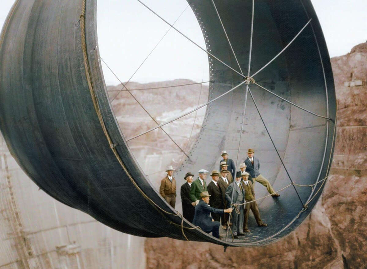 A group of men in suits and hats stand inside a large, curved metal pipe section suspended high above the ground, likely at a construction site in a rocky landscape.