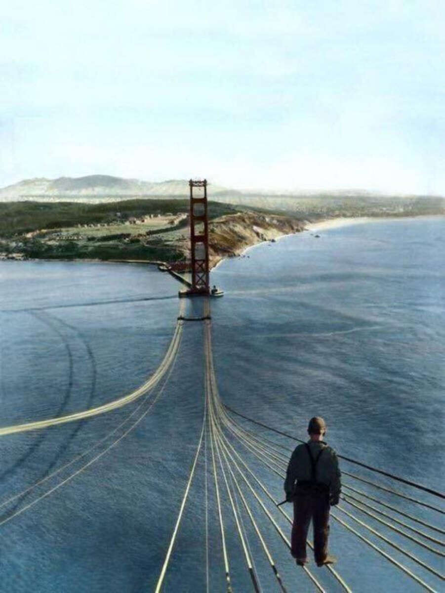A worker stands on suspension cables high above the water during the construction of the Golden Gate Bridge, with the partially built bridge and distant hills visible in the background.
