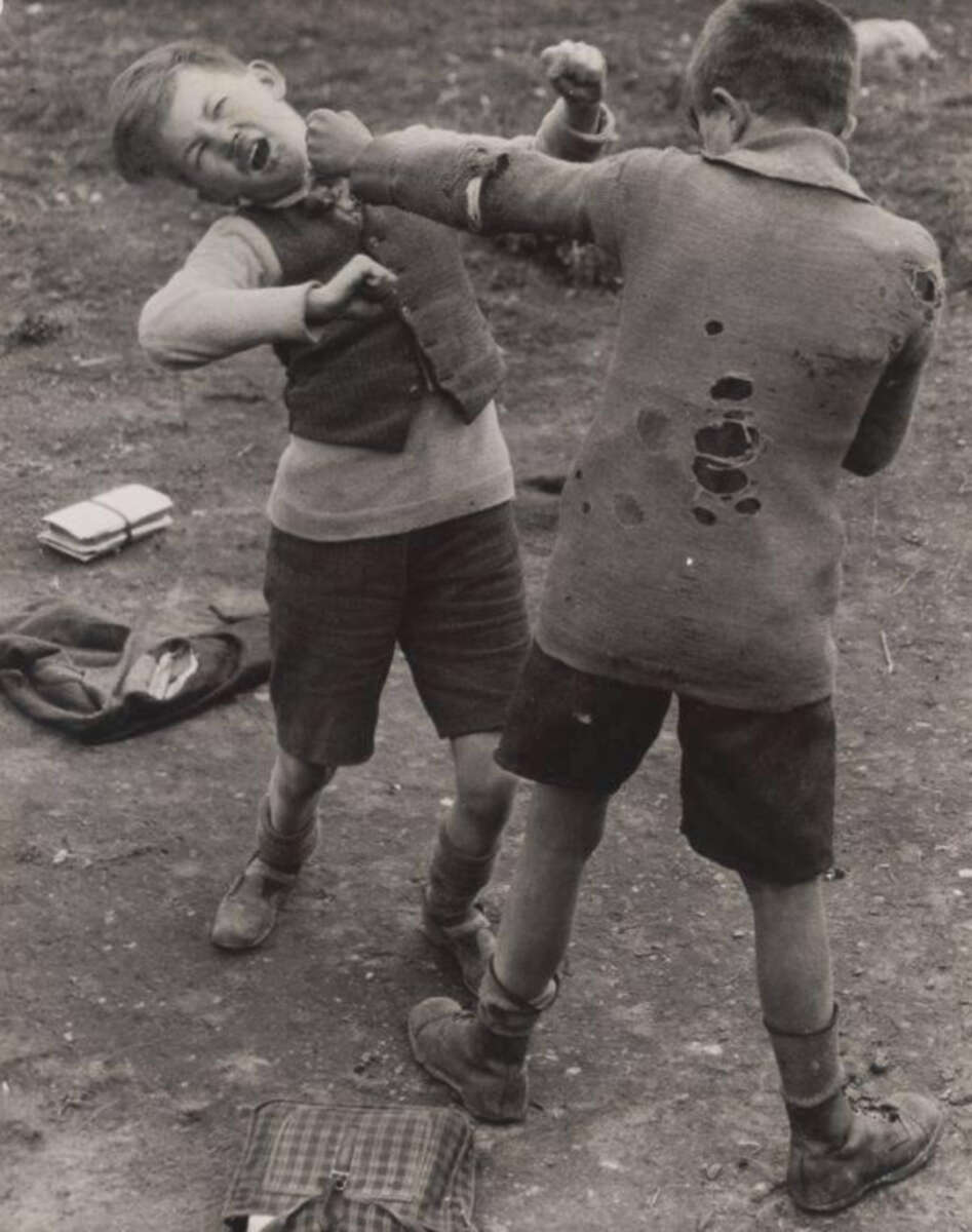 Two young boys in worn clothes are fighting outdoors; one boy punches the other, whose face shows pain. Schoolbags and jackets lie on the ground nearby. The scene suggests a rough, playful or serious quarrel.