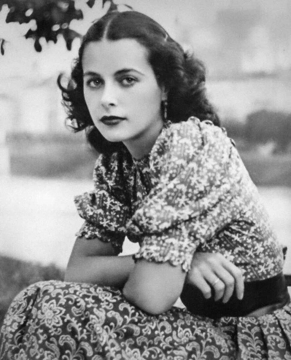 A woman with wavy dark hair and dark lipstick sits outdoors, wearing a patterned dress with puffed sleeves. She looks at the camera with a calm, serious expression. The background is blurred.