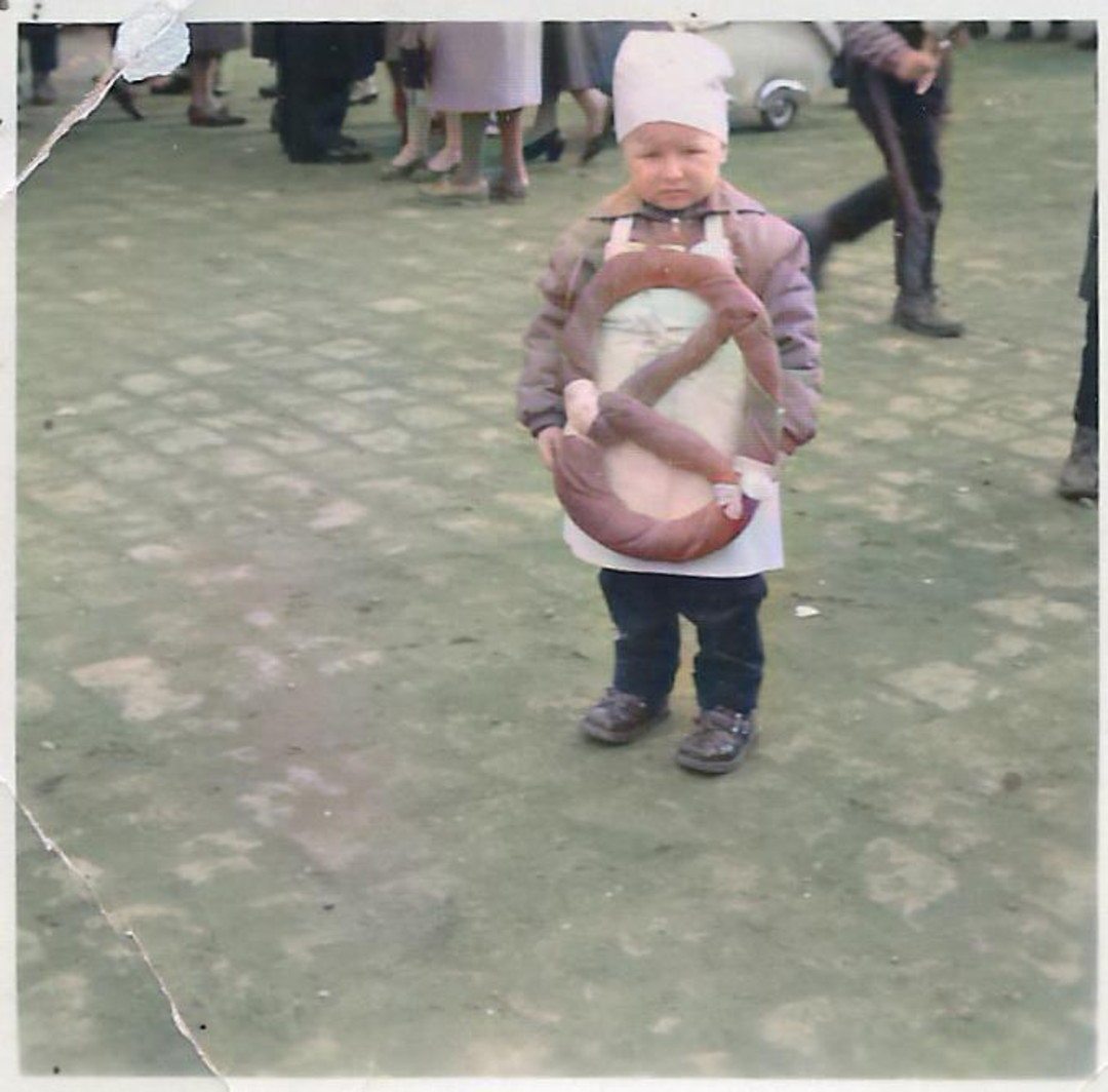 A young child wearing a beanie and jacket stands outdoors on a green, paved surface, holding a large sausage loop. Several blurred adults are visible in the background.