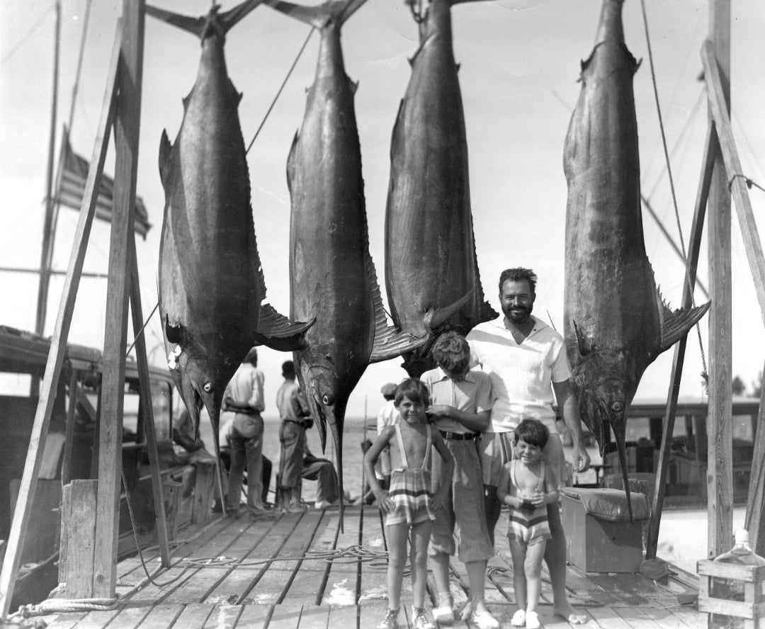 A man and four children stand on a dock beneath four large fish hanging vertically. Boats and an American flag are visible in the background, and the group appears to be smiling and enjoying the day.