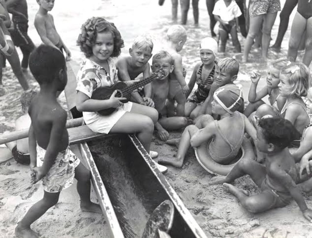A young girl sits on a canoe at the beach, playing a ukulele, surrounded by smiling children in swimsuits, some sitting in the sand and others standing, with the ocean in the background.