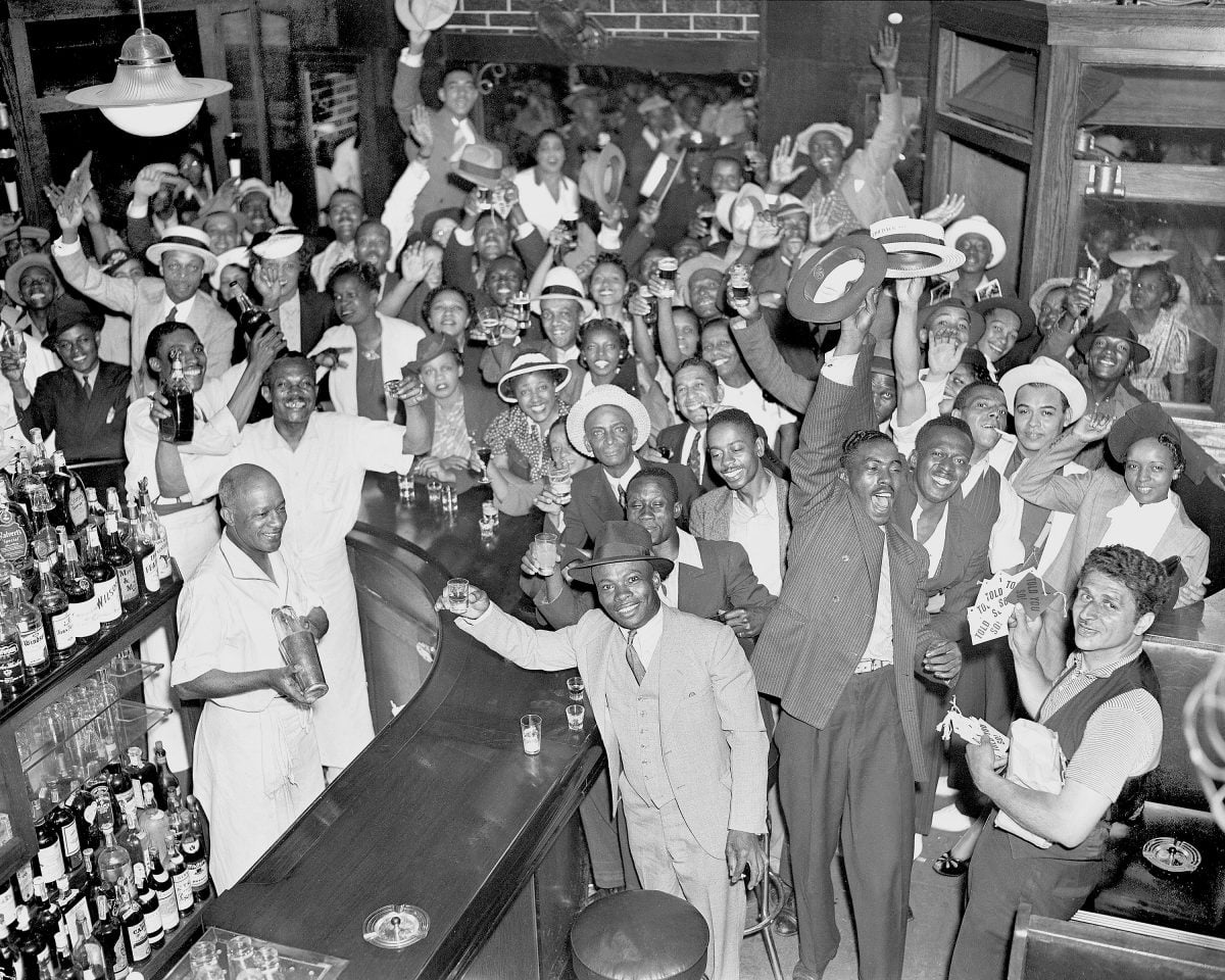A large group of people, mostly men, celebrate joyfully inside a crowded bar, many raising their hats and glasses. The bar is lively, with bartenders smiling and bottles lined up along the counter.