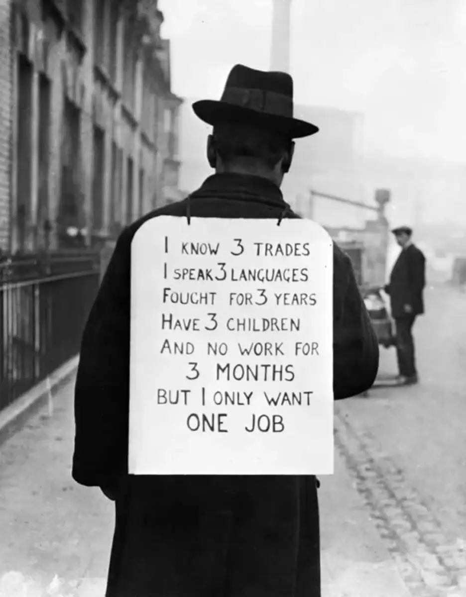 A man in a hat and coat stands on a street wearing a sign on his back that says he knows three trades, speaks three languages, fought for three years, has three children, and only wants one job after three months unemployed.