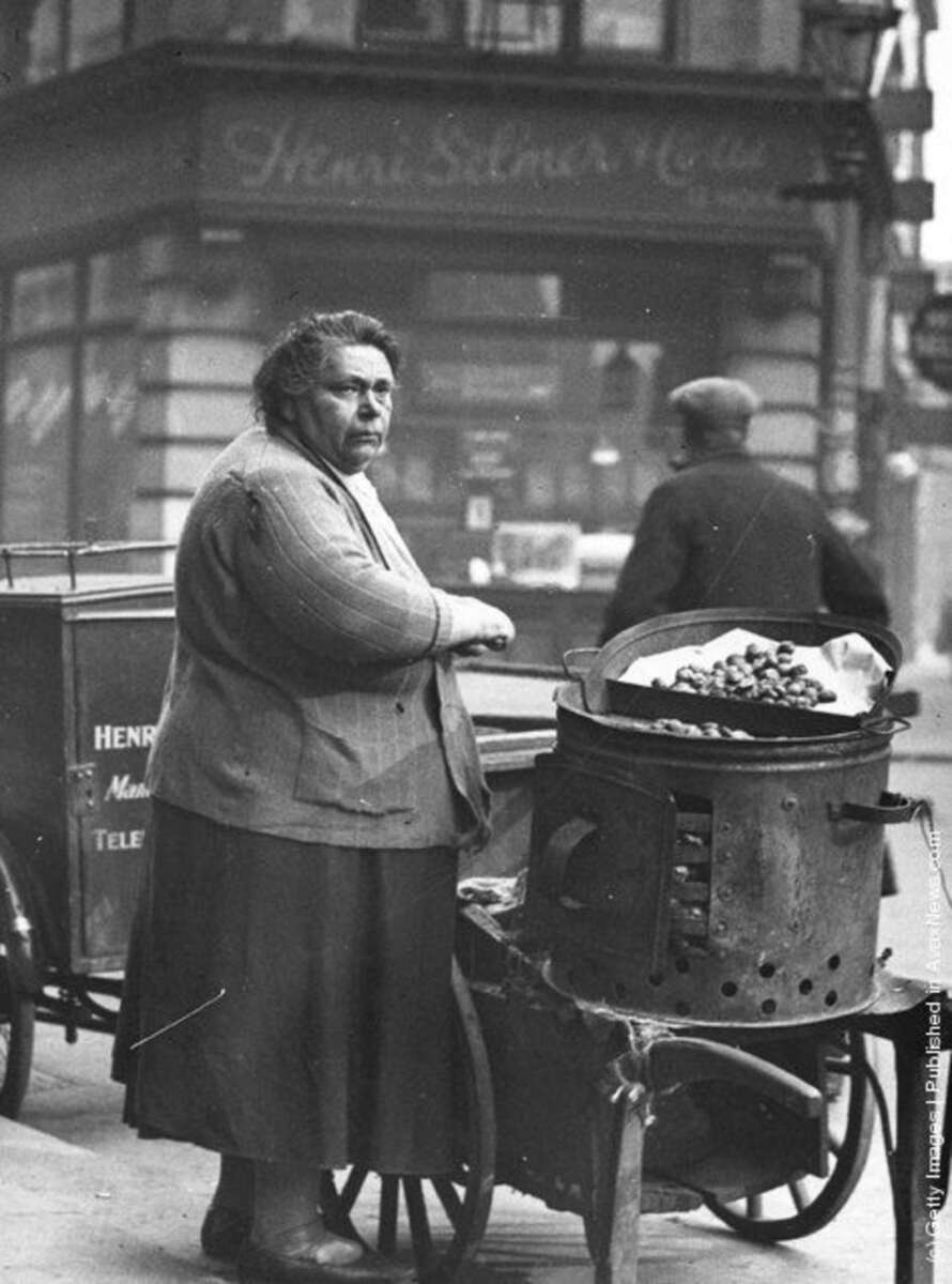 An older woman stands on a city sidewalk beside a cart with a metal drum roasting chestnuts. Another person walks by in the background near a storefront with French writing on the window.