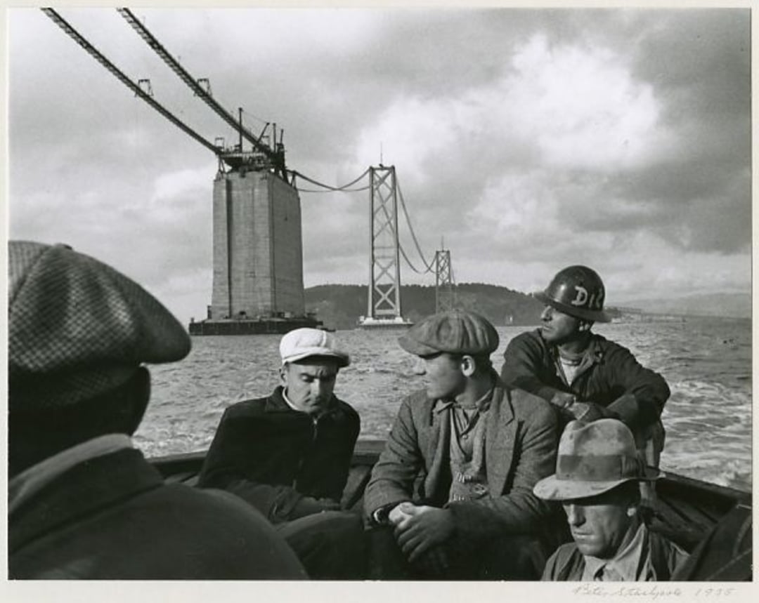 Four men in work attire sit in a boat on the water, with a suspension bridge under construction visible behind them. The sky is cloudy, and cables stretch across the incomplete bridge structure.