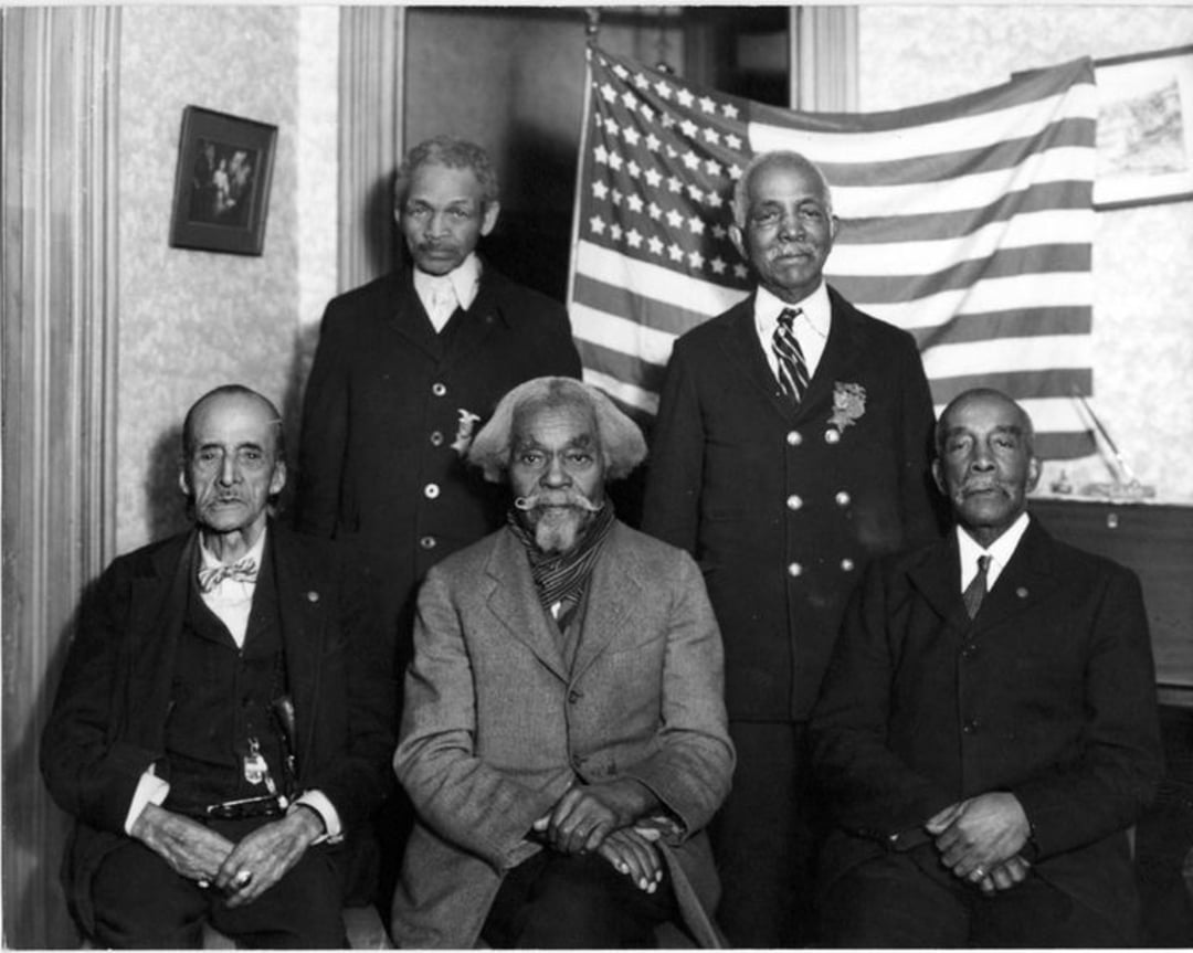Five older Black men in suits pose indoors, three seated and two standing, in front of a U.S. flag draped on the wall. A small framed picture hangs on the wall beside the flag.